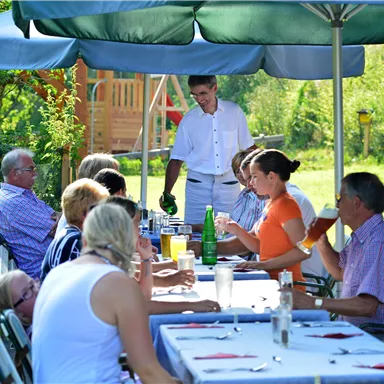 Eine Gruppe von Menschen sitzt an einem gedeckten Tisch im Freien. Ein Kellner serviert Getränke in einer schönen, grünen Umgebung.
