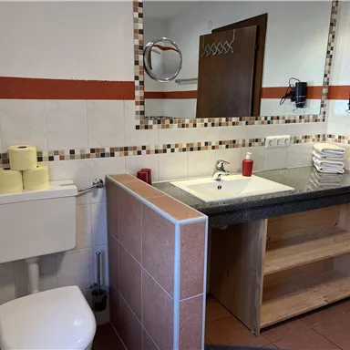 Ein modernes Badezimmer mit einem Waschbecken, Spiegel und Regalen. Es sind Toilettenpapierrollen und rote Kerzen dekorativ platziert.