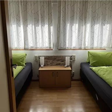 Ein einfaches Zimmer mit zwei Einzelbetten und hellen Vorhängen. In der Mitte steht ein kleiner Tisch mit zwei Lampen.