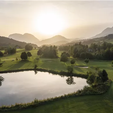 A picturesque landscape with a tranquil lake and gentle hills. The sun sets behind the mountains, casting warm light over the scene.