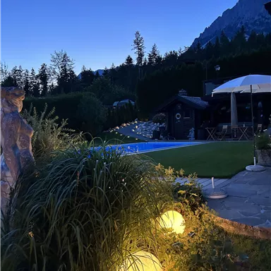 A beautiful garden with illuminated spheres and a pool. In the background, mountains and a cozy terrace can be seen.
