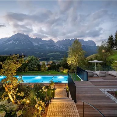 A modern terrace with a pool, surrounded by beautiful plants and a view of the mountains. The scene is lit at dusk and radiates a relaxed atmosphere.
