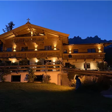 A cozy wooden house illuminated at night. In the background, you can see mountains and a clear evening sky.