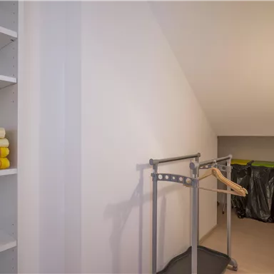 Ein heller Raum mit einem Kleiderständer und Regalen. Auf dem Regal liegen gelbe Handtücher, und im Hintergrund sind einige abgedeckte Gegenstände zu sehen.