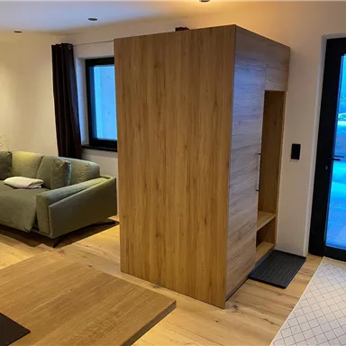 Ein moderner Wohnraum mit einem Holzwürfel als Raumtrenner. Im Hintergrund sind ein Sofa und große Fenster mit Blick nach draußen zu sehen.