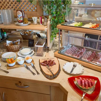 Ein Buffet mit verschiedenen Speisen, darunter Aufschnitt, Käse und Obst. Auf dem Tisch liegen zusätzlich Teller und Gläser.