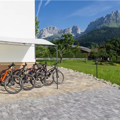 Eine Gruppe von Fahrrädern steht unter einem Vordach auf einem Pflasterplatz. Im Hintergrund sind Berge und eine grüne Wiese zu sehen.