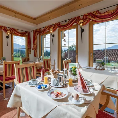 Ein einladender Frühstücksraum mit hellen Fenstern und schöner Aussicht. Auf den Tischen sind verschiedene Frühstücksgerichte und Getränke angerichtet.