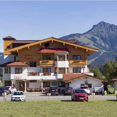 Ein charmantes Hotel mit Holzelementen, umgeben von grünen Wiesen und Bergen. Der Himmel ist klar und sonnig, was eine einladende Atmosphäre schafft.