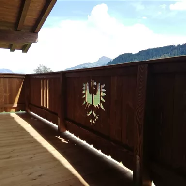 Eine Holzveranda mit einem dekorativen Geländer und Blick auf die Berge. Der Himmel ist klar und es ist sonnig.
