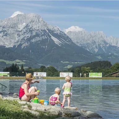 Eine Mutter sitzt am Ufer eines Sees mit ihren zwei Kindern. Im Hintergrund sind majestätische Berge und ein klarer blauer Himmel zu sehen.