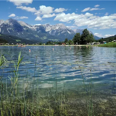 A clear lake surrounded by meadows and mountains. The sky is blue and adorned with some clouds.