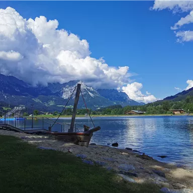 A quiet lake surrounded by mountains and green meadows. The clouds are big and white against a clear blue sky.