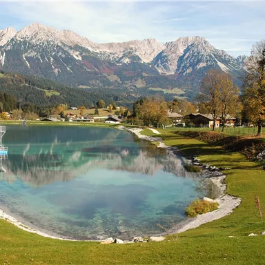 A picturesque view of a clear lake, surrounded by green meadows and majestic mountains. The calm water surface reflects the surrounding nature.