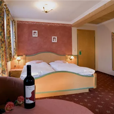 Ein gemütliches Schlafzimmer mit einem Doppelbett und warmer Farbgestaltung. Auf einem Tisch steht eine Flasche Wein und eine Rose.