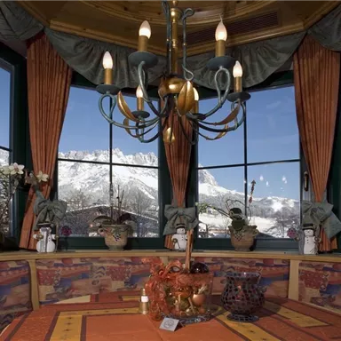 Ein gemütlicher Raum mit Fenstern, die einen Blick auf schneebedeckte Berge bieten. Die Einrichtung ist rustikal mit Pflanzen und einem Kronleuchter.