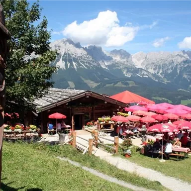A cozy mountain cabin with colorful umbrellas outside. In the background, majestic mountains rise under a blue sky.