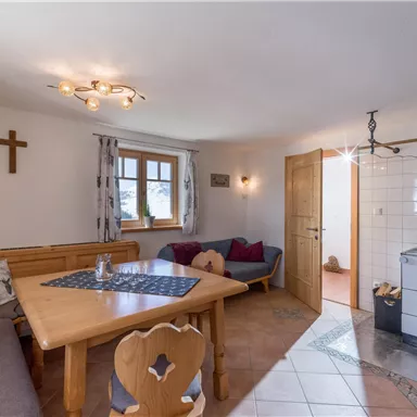 Eine gemütliche Wohnküche mit einem großen Tisch, einer Sitzecke und einem Holzofen. Helle Wände und Fenster sorgen für eine einladende Atmosphäre.