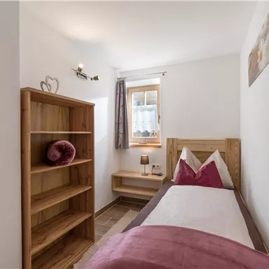 Ein einfaches Schlafzimmer mit einem Einzelbett und einer Holzregal. Die Wände sind hell und es gibt ein Fenster mit Vorhängen.