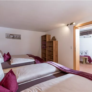 Ein gemütliches Schlafzimmer mit zwei Betten und modernen Holzelementen. Im Hintergrund befindet sich ein Regal und ein Zugang zu einem weiteren Raum.