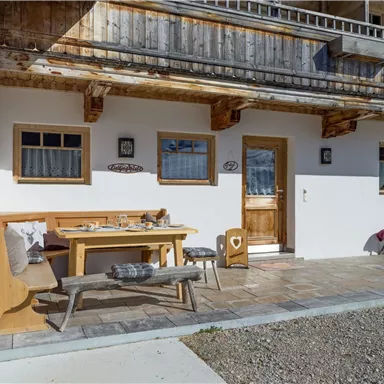 Eine gemütliche Terrasse mit einem Holztisch und Bänken. Im Hintergrund sind traditionelle Holzelemente und Fenster zu sehen.