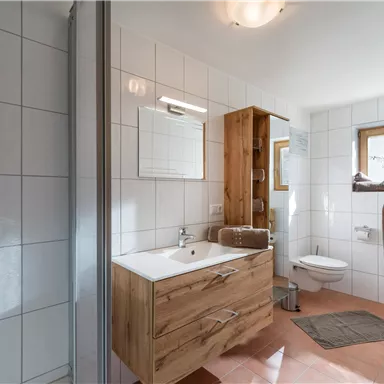 Ein modernes Badezimmer mit einer großzügigen Dusche und Holzschrank. Helle Fliesen und große Fenster sorgen für eine einladende Atmosphäre.