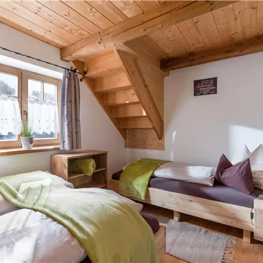 Ein gemütliches Schlafzimmer mit Holzdecke und zwei Betten. Ein Fenster mit Ausblick und Treppe verleihen dem Raum eine helle, einladende Atmosphäre.