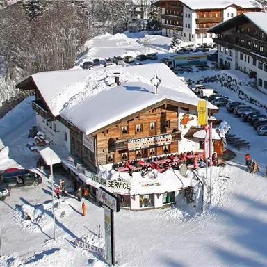 Ein gemütliches Holzhaus in einer verschneiten Umgebung. Das Gebäude ist von Schnee umgeben und es sind viele Skifahrer in der Nähe.