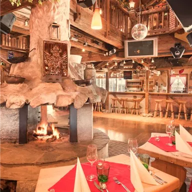 Ein gemütliches Restaurant mit einem offenen Kamin und rustikalen Holzmöbeln. Die Tische sind liebevoll gedeckt und es strahlt eine einladende Atmosphäre aus.