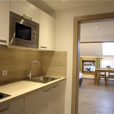 A modern kitchen with wooden surfaces and a microwave. In the background, a dining area with a table and chairs is visible.