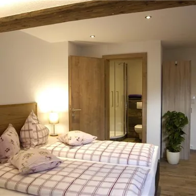 A cozy bedroom with two single beds and checkered pillows. The room is bright and modernly furnished, with a bathroom nearby.