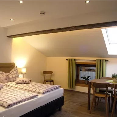 A cozy bedroom with a double bed and comfortable pillows. There is a small dining area with a table and chairs as well as a window with green curtains.