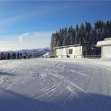 A snowy mountain landscape with a long line of skiers. In the background, there are trees and a blue sky.