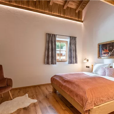 Ein modernes Schlafzimmer mit einem Doppelbett und Holzdekor. Die Wände sind hell und es gibt einen Fernseher sowie einen gemütlichen Sessel.