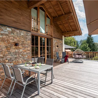 Eine einladende Terrasse mit einem Holztisch und Stühlen. Im Hintergrund ist ein modernes Holzhaus und schöne grüne Bäume zu sehen.