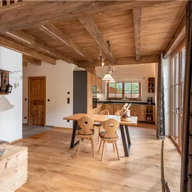 Eine gemütliche Küche mit Holzmöbeln und großen Fenstern, die viel Licht hereinlassen. Der Boden ist aus Holz, und es gibt einen einladenden Esstisch mit Stühlen.