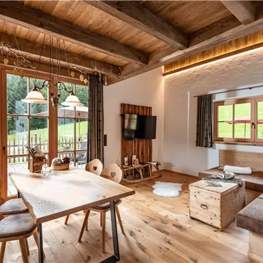 Ein modernes, gemütliches Wohnzimmer mit einem großen Esstisch und Holzdekor. Durch die Fenster ist die grüne Landschaft sichtbar, die viel Licht hereinlässt.