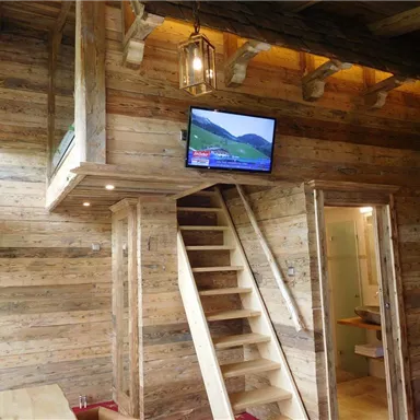 Ein gemütlicher Innenraum aus Holz mit einer Treppe, die zu einer oberen Etage führt. An der Wand hängt ein Fernseher, und das Licht wird durch rustikale Lampen hervorgehoben.