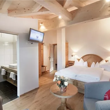 A modern room with a double bed and light wooden furniture. In the background, a bathroom and a television can be seen.