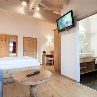 A modern room with a double bed, a television, and a cozy seating area. The bathroom is visible through a glass wall, designed elegantly and brightly.