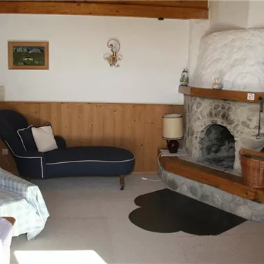 Ein gemütlicher Wohnraum mit einem Steinofen und einer blauen Couch. An der Wand hängen Bilder, und es gibt einen Holztisch mit Stühlen.