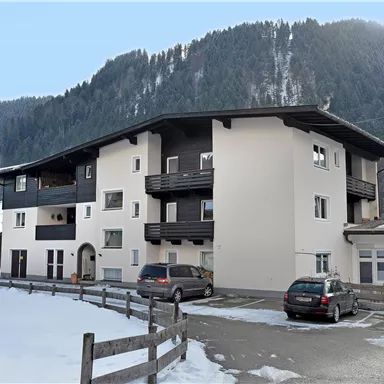 A modern residential building in a snow-covered area. In the background, mountains and a clear sky can be seen.
