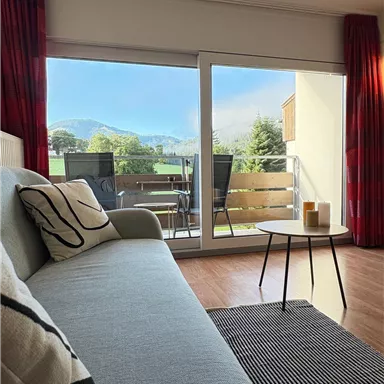 A cozy living space with a bright sofa and a view of the mountains. Outside is a balcony with garden furniture and a beautiful view.