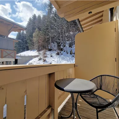 Ein Balkon mit einem kleinen Tisch und einem Stuhl, umgeben von schneebedeckten Bäumen. Der Himmel ist klar und blau, was eine schöne Aussicht bietet.
