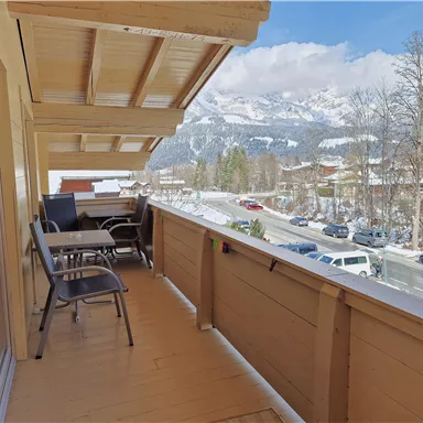 Ein Balkon mit Holzverkleidung, auf dem Stühle und ein Tisch stehen. Im Hintergrund sind schneebedeckte Berge und eine klare blaue Himmel zu sehen.
