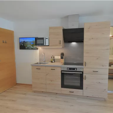 Eine moderne Küchenzeile aus Holz mit einem Herd, einem Kühlschrank und einer Mikrowelle. Im Hintergrund ist ein Badezimmer zu sehen, und es gibt einen Fernseher an der Wand.