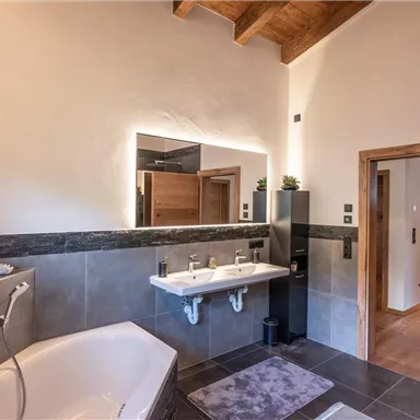 Ein modernes Badezimmer mit einer Badewanne und einem Doppelwaschbecken. Es hat helle Wände, dunkle Fliesen und natürliche Holzelemente.