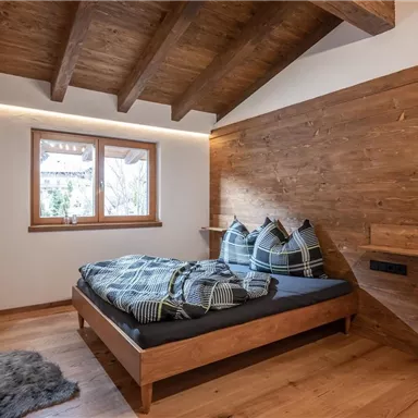 Ein modernes Schlafzimmer mit einem bequemen Bett und einem Holzwandpaneel. Große Fenster sorgen für viel Tageslicht und eine gemütliche Atmosphäre.