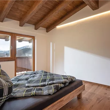 Ein gemütliches Schlafzimmer mit einem Holzbett und karierten Decken. Große Fenster lassen viel Tageslicht und einen Blick auf die Berge herein.