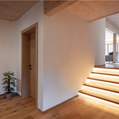 Ein modernes Treppenhaus mit Holztreppen und sanfter Beleuchtung. Die Wände sind hell gestrichen, und eine Pflanze steht in der Ecke.